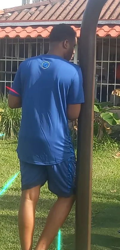 Man in blue outfit leaning on pole in sunny garden.