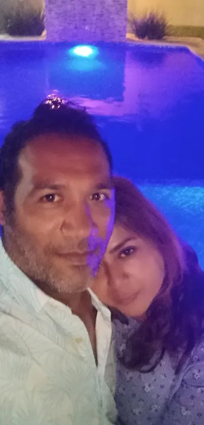 Couple enjoying a nighttime selfie by a blue-lit pool.