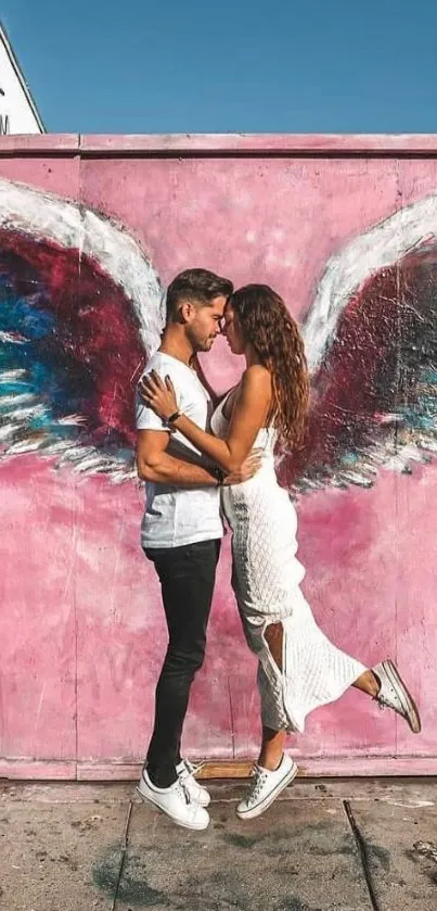 Couple embraces in front of colorful street art with pink wings.