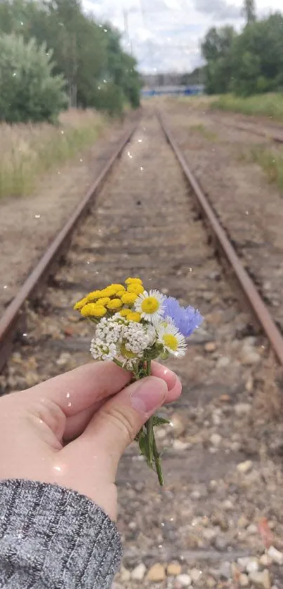 Hand holding wildflowers by railroad tracks.