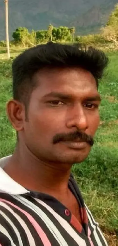 Man in a striped shirt standing in a lush green field with hills in the background.