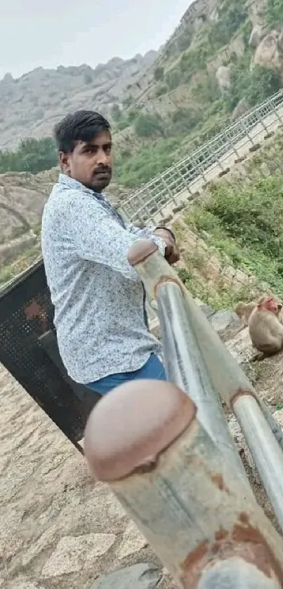 Man stands by a railing, overlooking a scenic hilltop view with greenery.