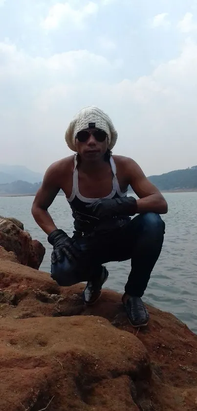 Person squatting on rocks by serene lake with mountains, wearing white beanie.