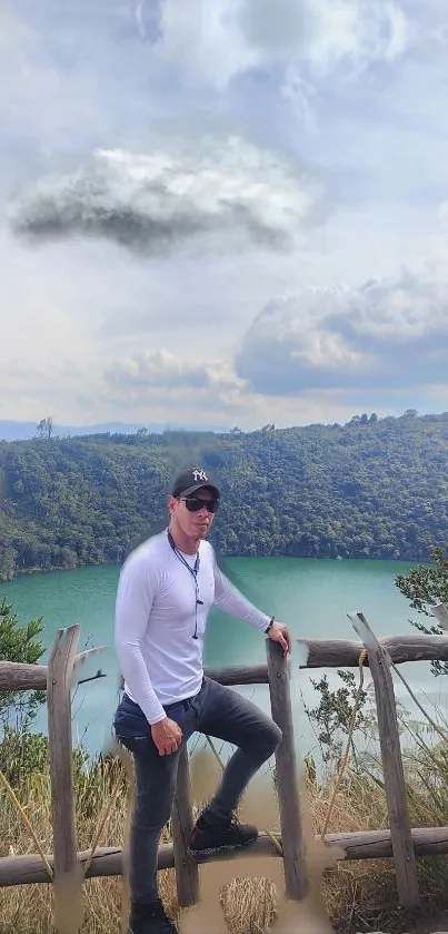 A person stands by a wooden fence overlooking a serene lake with hills and a cloudy sky.