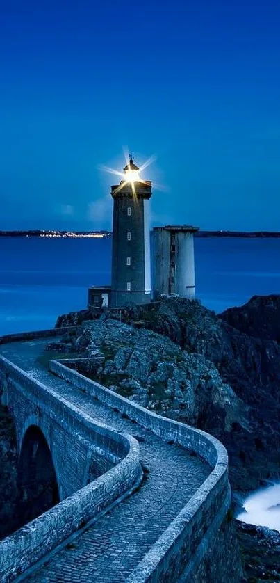 Lighthouse glowing at night on rocky cliffs with a blue ocean backdrop.