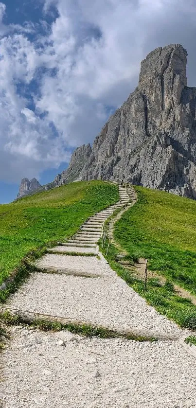 Pathway leading up a green hill toward a rocky mountain under a blue sky.