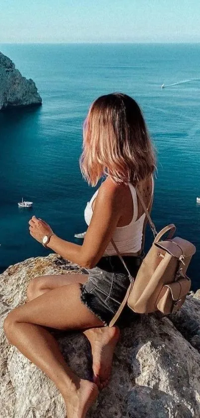 Woman seated on cliff overlooking calm blue ocean with distant sailboats.