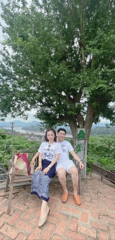 Couple sitting on a bench under a lush green tree in a scenic outdoor setting.