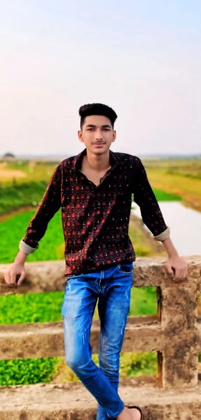 Young man on bridge with green fields and river backdrop.