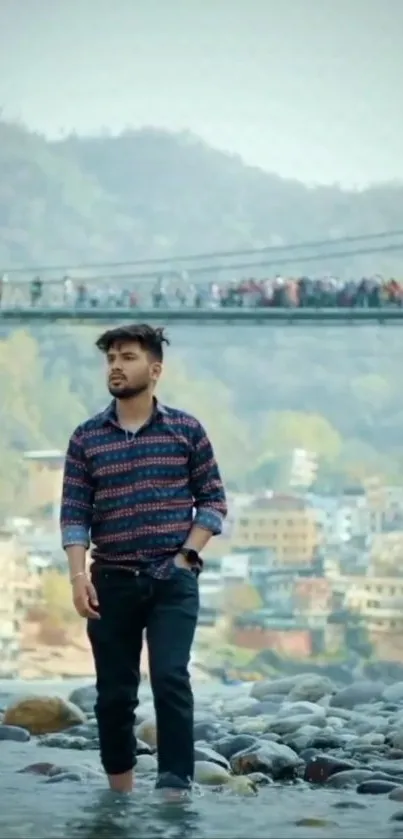 Man walking in scenic riverside with bridge and mountain backdrop.