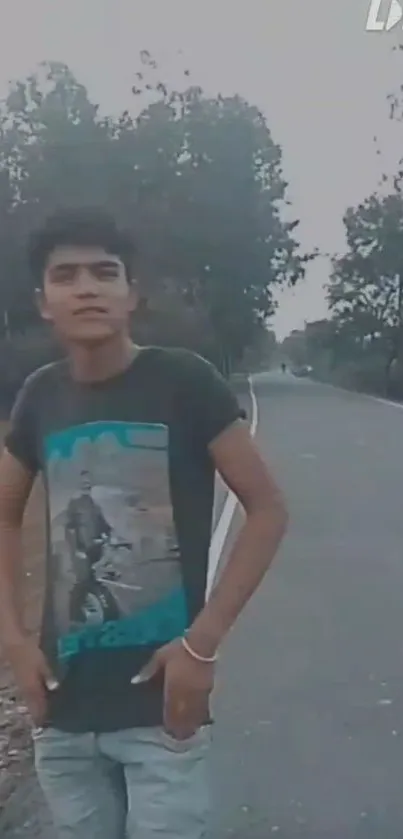 Young man standing on a scenic roadside with lush trees and a peaceful road.