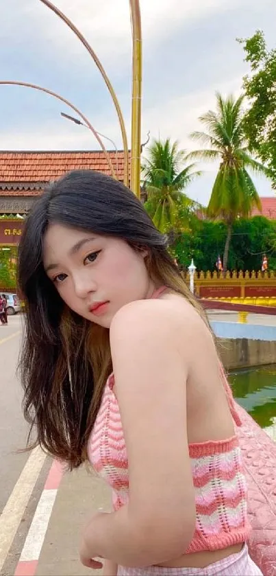 Young woman in pink top by a scenic Asian street with trees and traditional architecture.