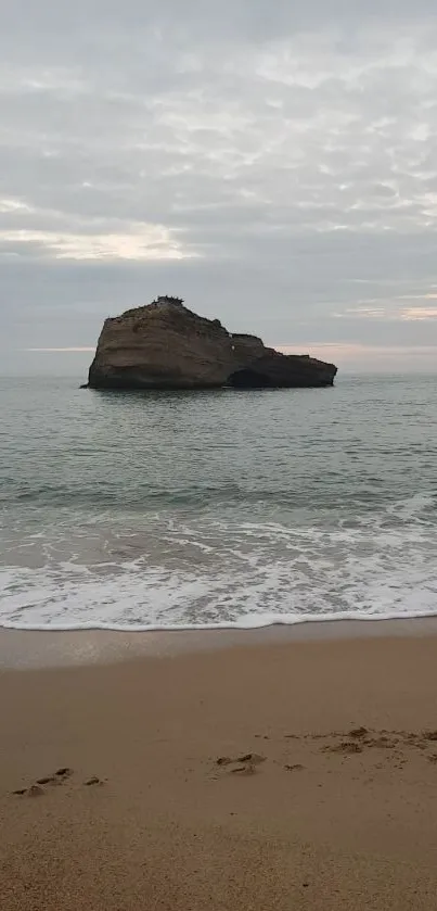 Tranquil beach with ocean rock under cloudy sky.