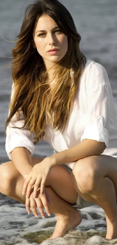 Woman crouching by the beach, wearing white blouse and shorts.