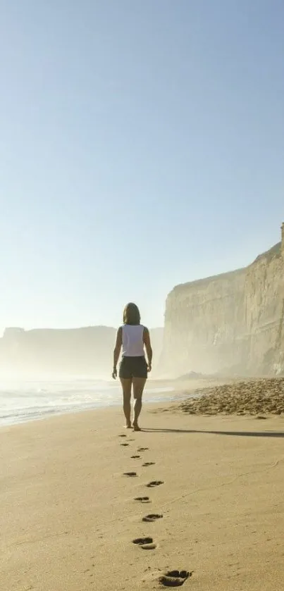 Person walking along a sunlit beach with cliffs and footprints in the sand.