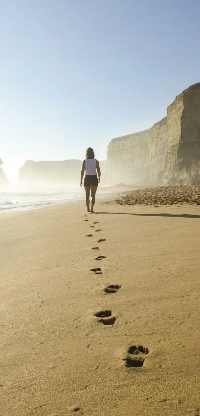 A person walking on a beach, leaving footprints in the sand under a clear blue sky.