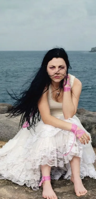 Woman in white dress sitting on rocks by the ocean with a serene expression.