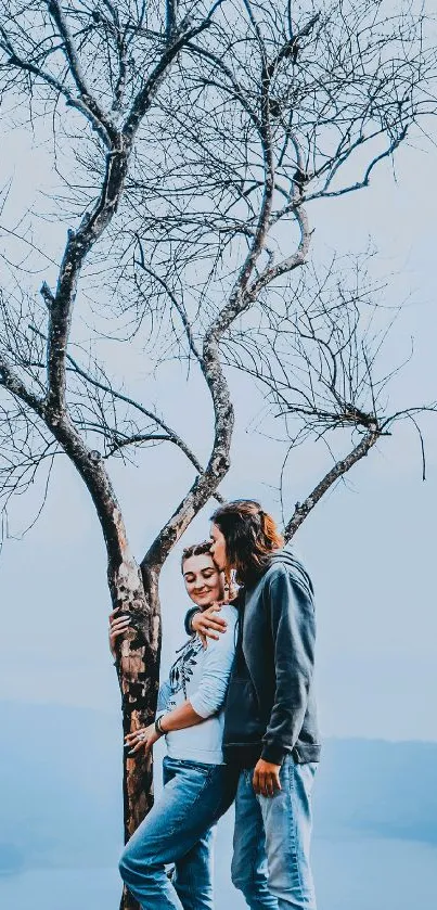 Couple embracing under a bare tree with a blue sky backdrop.