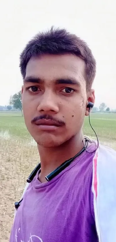 Young man in purple shirt with earphones standing in serene field.