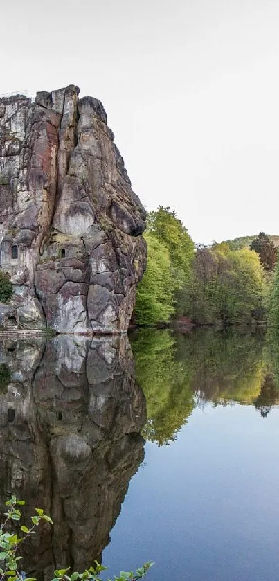 Towering cliff reflects in calm lake with lush, green trees around.