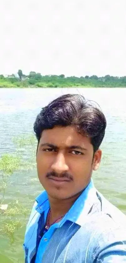 Man taking a selfie by a green lakeside with clear blue sky.