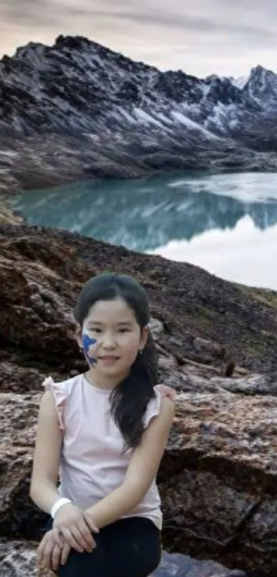 Young girl sitting by a mountain lake under a scenic sky.