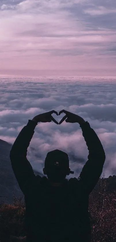 Silhouette of person forming heart with hands against sunset over mountains and clouds.