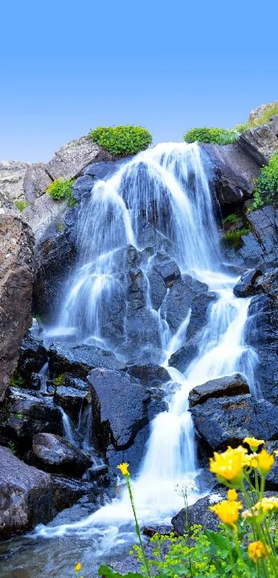 Serene waterfall on rocky mountain with wildflowers under a blue sky.