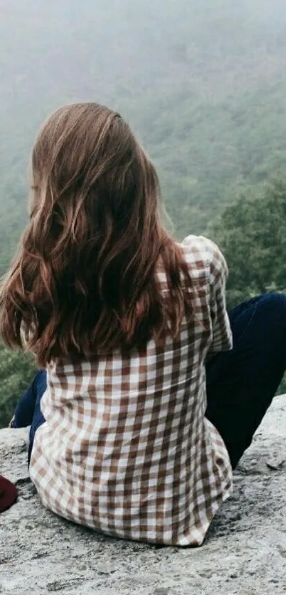 Woman in plaid shirt sitting on a rock, overlooking misty forest landscape.