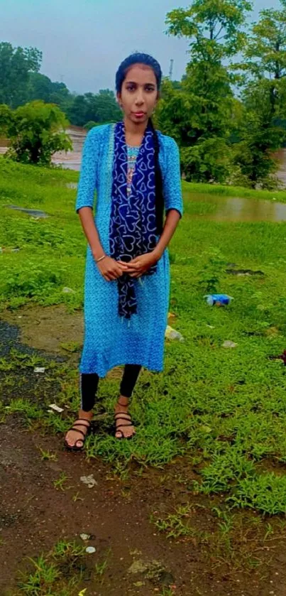 Woman standing outdoors in blue dress with green landscape background.