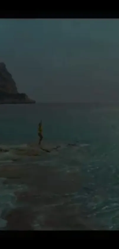 Person on rock by the serene teal ocean with mountain backdrop.