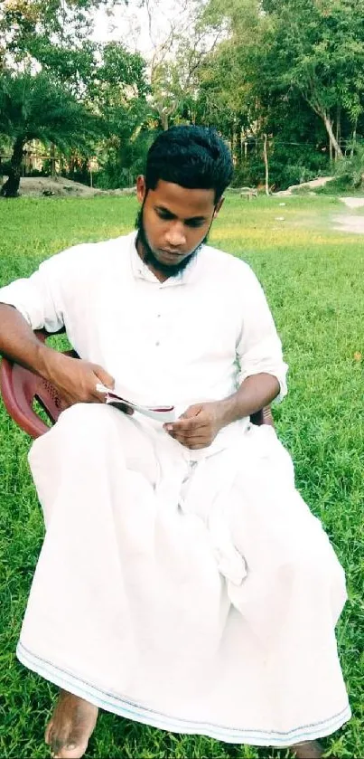 A person in white clothing sitting outdoors on grass, reading.