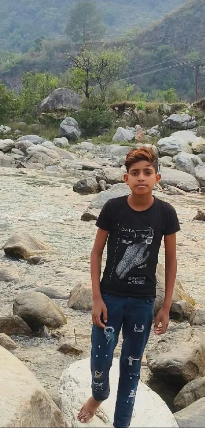 Young boy stands by a rocky river in a serene mountain landscape.