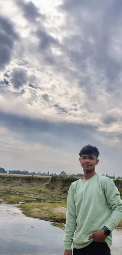 Young man stands by a river under a cloudy sky.