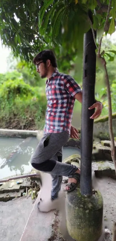 Man in plaid shirt relaxes by river under lush greenery.