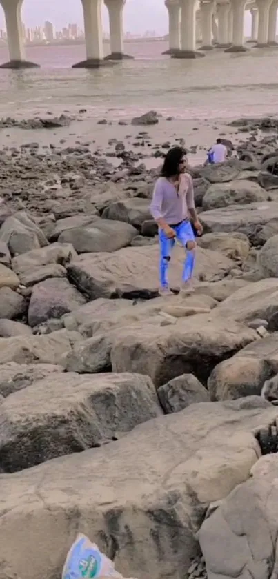 Woman walking along a rocky beach with a city view.