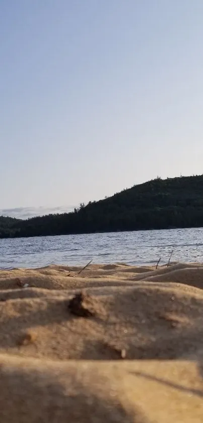 Sandy beach with lake and forested hills under clear sky.