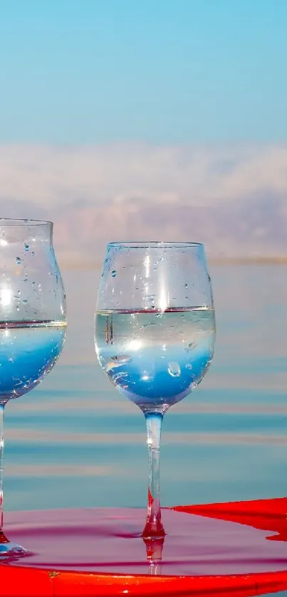 Two wine glasses on a tray in a serene sea setting with blue sky and mountains.