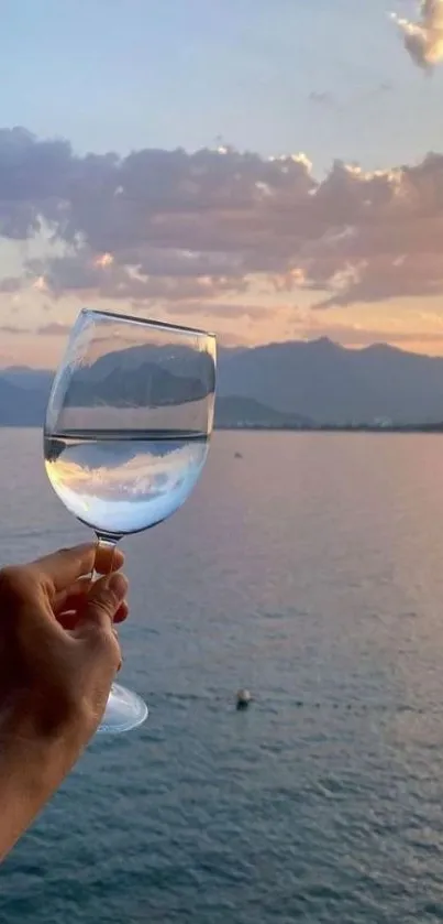 Hand holding glass with serene sunset view over ocean.