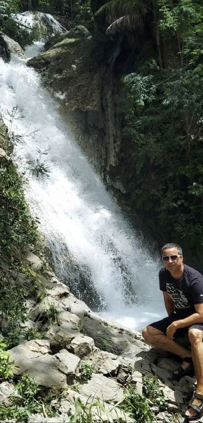 Man sitting near a lush waterfall with greenery surrounding.