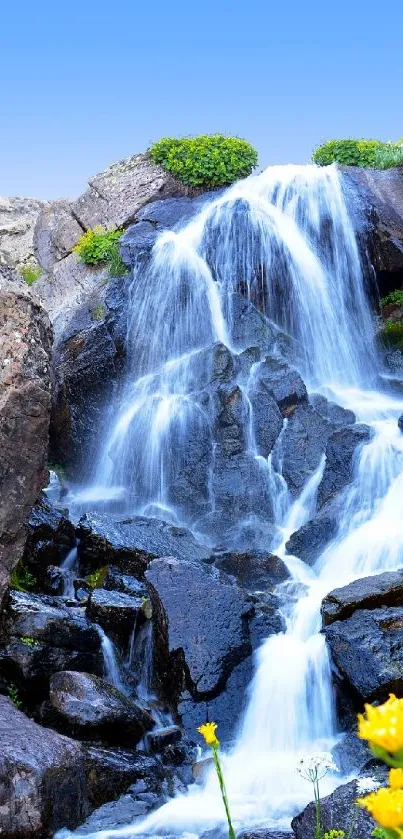 Serene waterfall cascading over rocks with a clear blue sky and wildflowers.