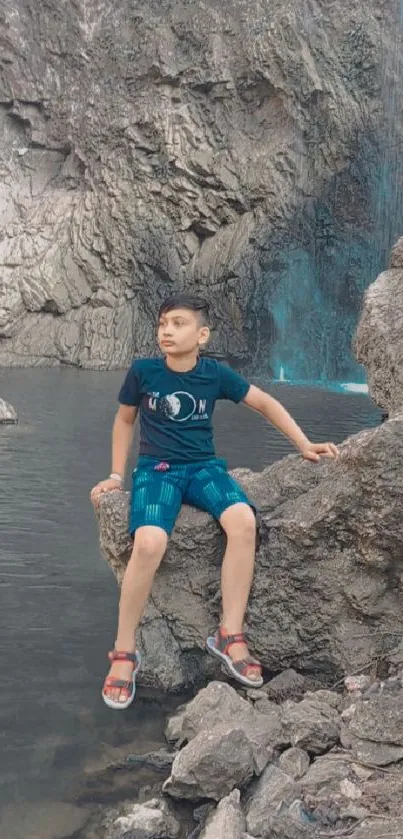 Boy sitting on rocks by a serene waterfall amidst nature.