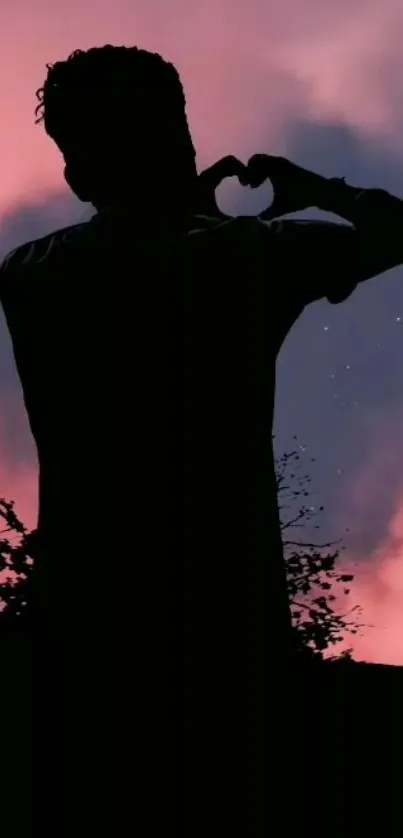 Silhouette of person against pink sunset sky making a heart shape.