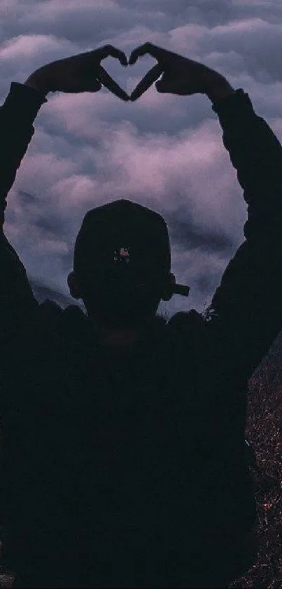 Silhouette of person forming heart hands in cloudy sky.