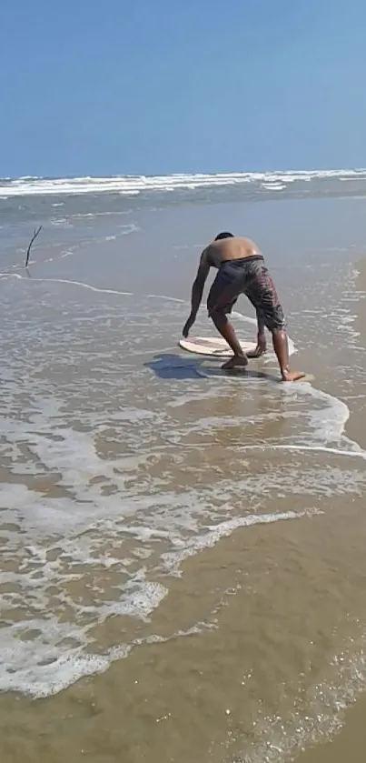 Person skimboarding on a sunny beach with waves and clear blue sky.