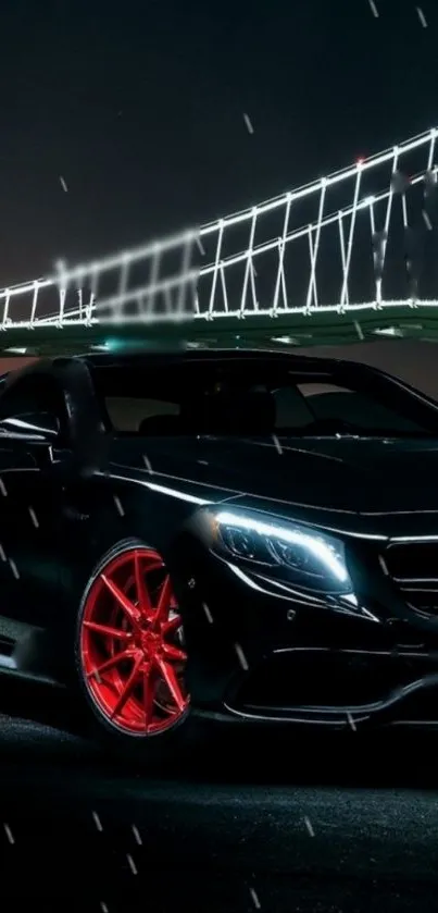 Sleek black car with red rims under a lit bridge at night.