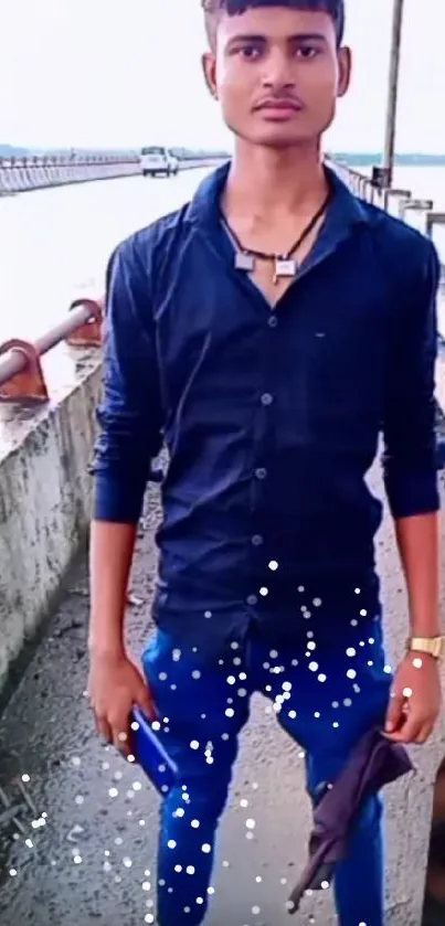 Young man in blue shirt posing outdoors by a bridge.