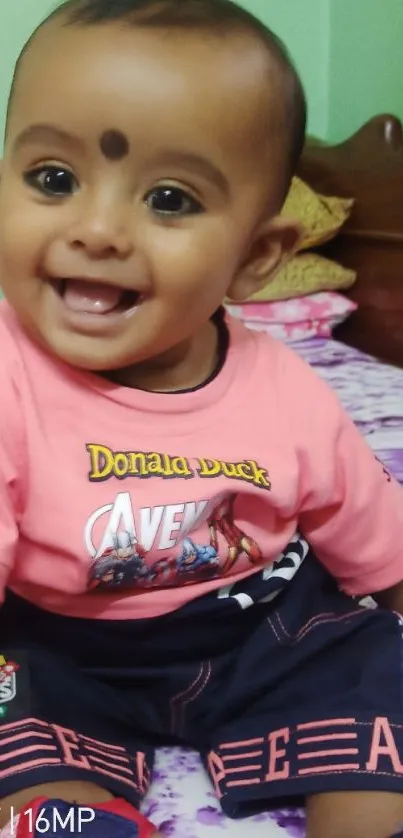 Smiling baby sitting on a bed with a floral bedspread wearing a pink outfit.