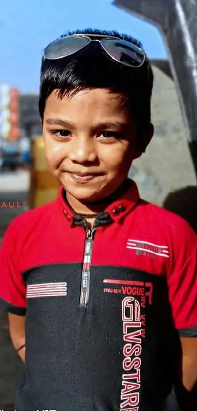 A young boy smiles in a red shirt with sunglasses, set against a bright urban backdrop.