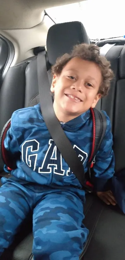 A smiling child in blue outfit sits in car seat, secured by a seatbelt.